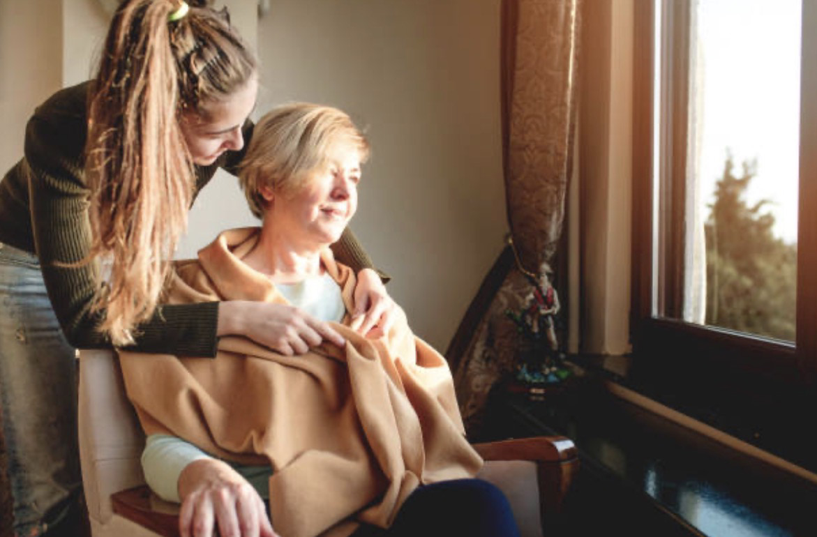 A care professional supporting an older woman seated beside a window